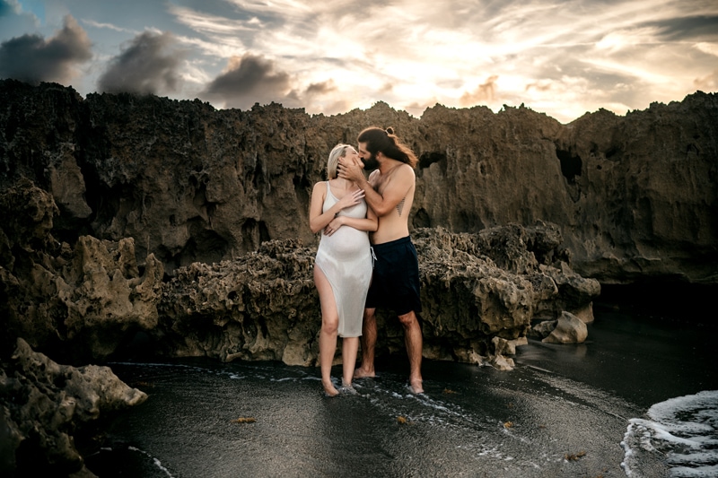 Maternity Photographer, man and expecting woman stand in shallow waters at beach near rocks